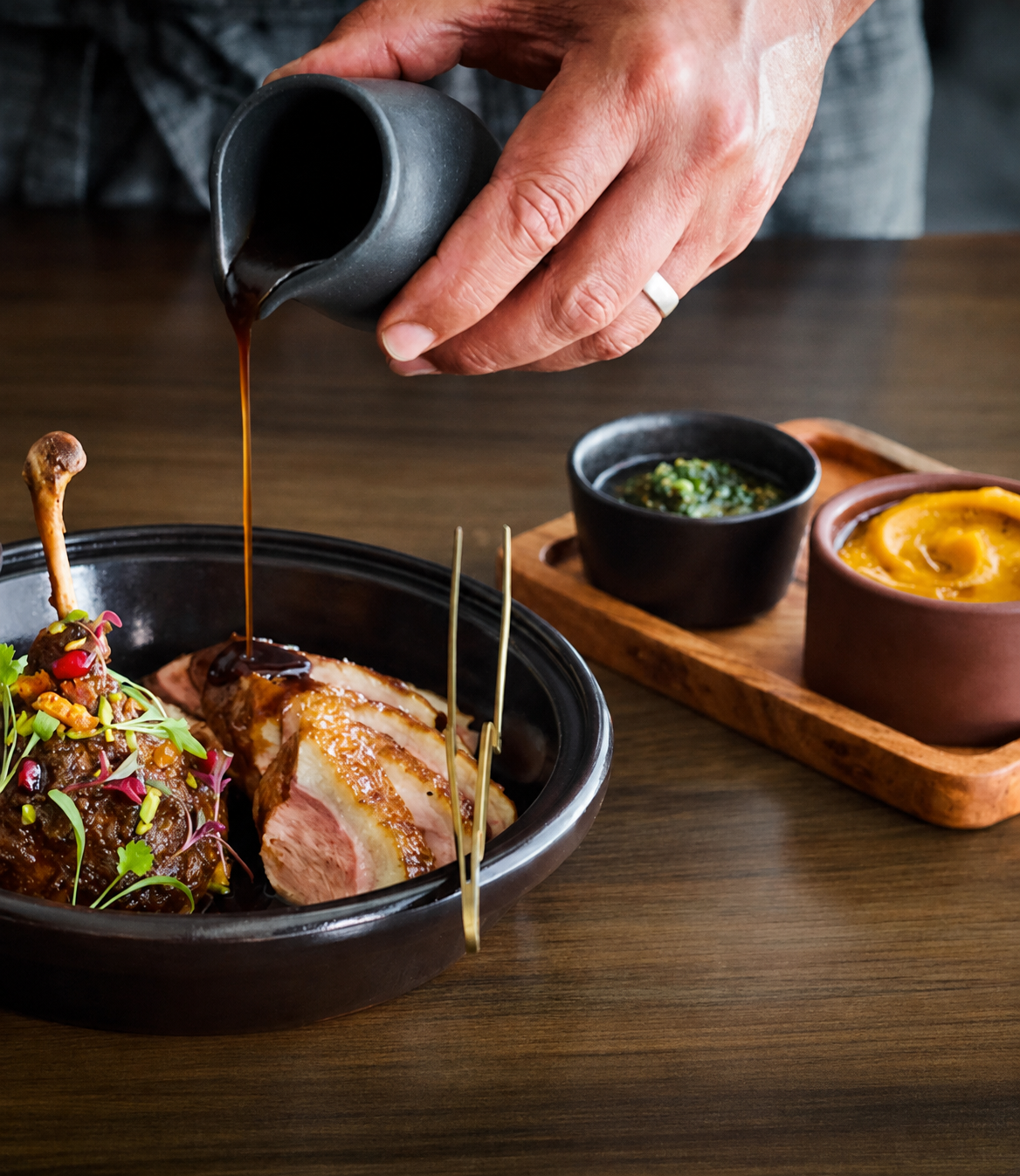 Chef pouring rich sauce over sliced roasted meat served with vegetables and colourful side dishes.