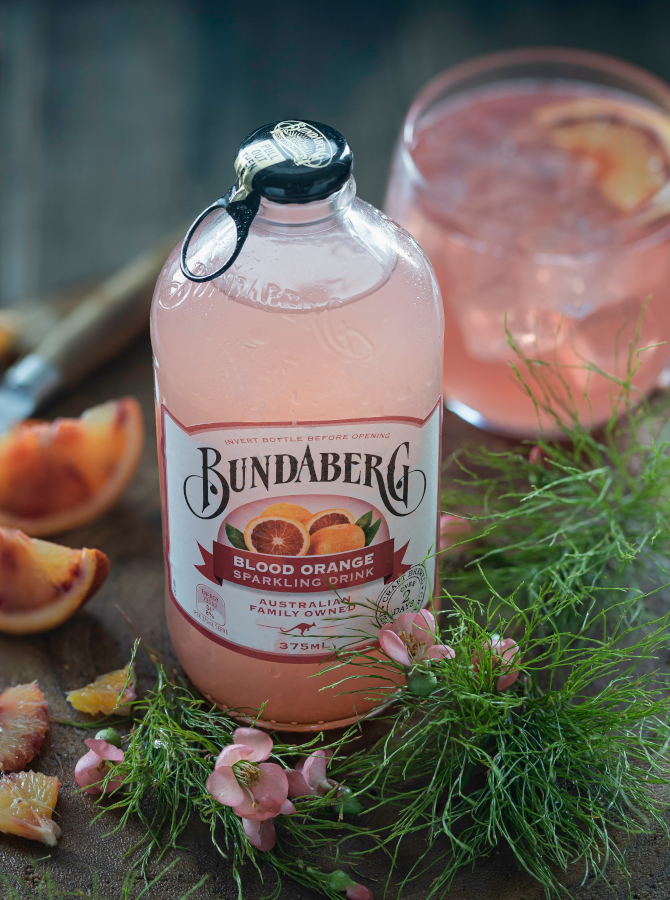 Bundaberg Blood Orange sparkling drink bottle surrounded by fresh citrus, greenery and a pink drink.